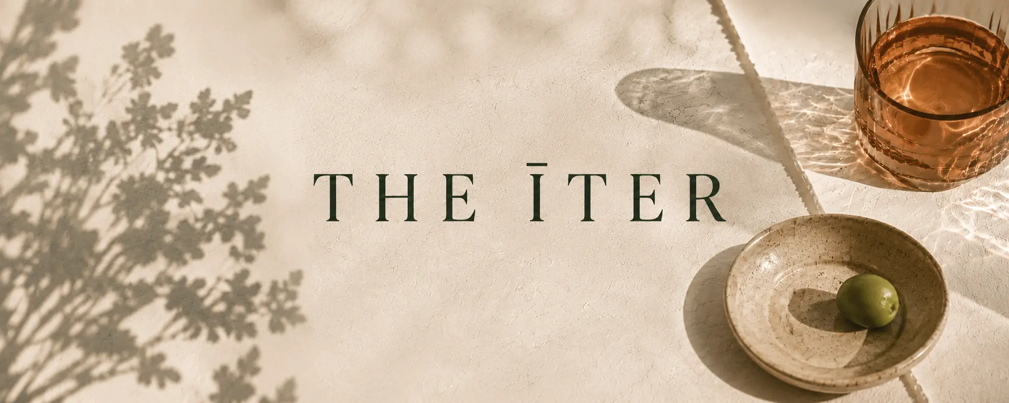THE ĪTER on a warm still life of glass, olive, and botanical shadows